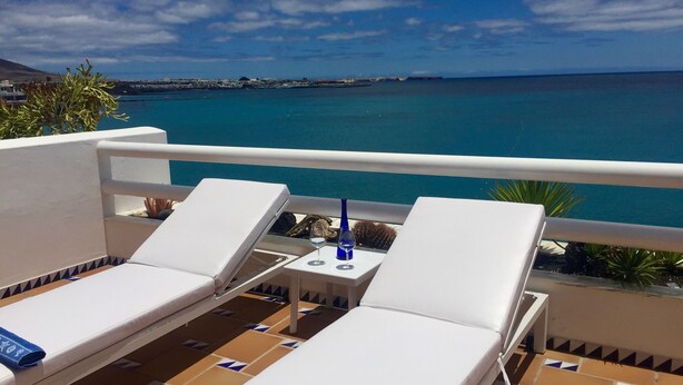 Een balkon met twee witte lounge fauteuils, een klein tafeltje met een glas wijn, en uitzicht op zee en verre gebouwen.