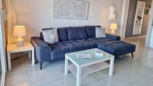 Een moderne woonkamer met een grijze modulaire bank, twee bijzettafels met lampen, een glazen salontafel en een abstract kunstwerk aan de muur.