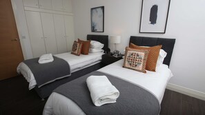 A hotel room with two beds, each with a grey bedspread and white linens, a bedside table with a lamp, and a wardrobe.