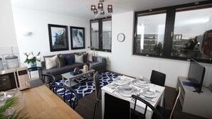 A modern kitchen with a microwave, kettle, and a coffee maker. There is a dining table set with plates, cutlery, and a vase with flowers. A sofa with cushions is against the wall, and there are two framed pictures above it.