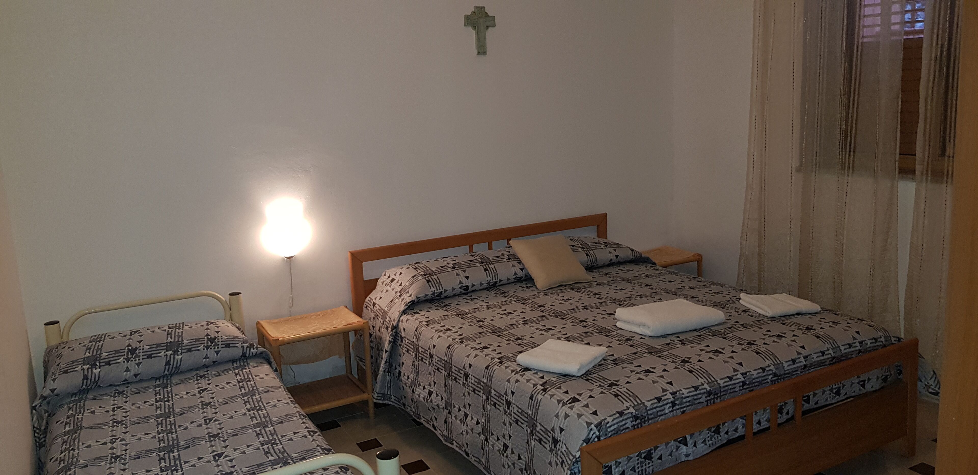 Une chambre avec deux lits, une table de chevet, une lampe fixée au mur et une fenêtre avec des rideaux.