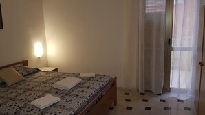 Een net opgemaakt bed met een beddekleed met patroon en kussens, een nachtkastje met een lamp, en uitzicht naar buiten door een raam met dunne gordijnen.