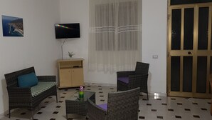 Een woonkamer met een bank, twee fauteuils, een salontafel, een televisie, een kast, een raam met kantgordijnen, en een deur.