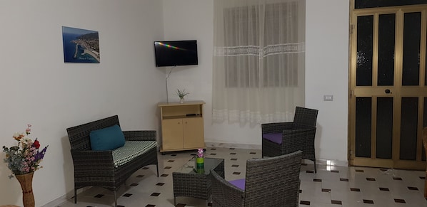 A living room with a sofa, two armchairs, a coffee table, a TV, a cabinet, a window with lace curtains, and a door.