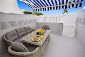 Une terrasse avec une table dressée pour le petit-déjeuner, comprenant des croissants, des fruits et du jus, sous une marquise à rayures.