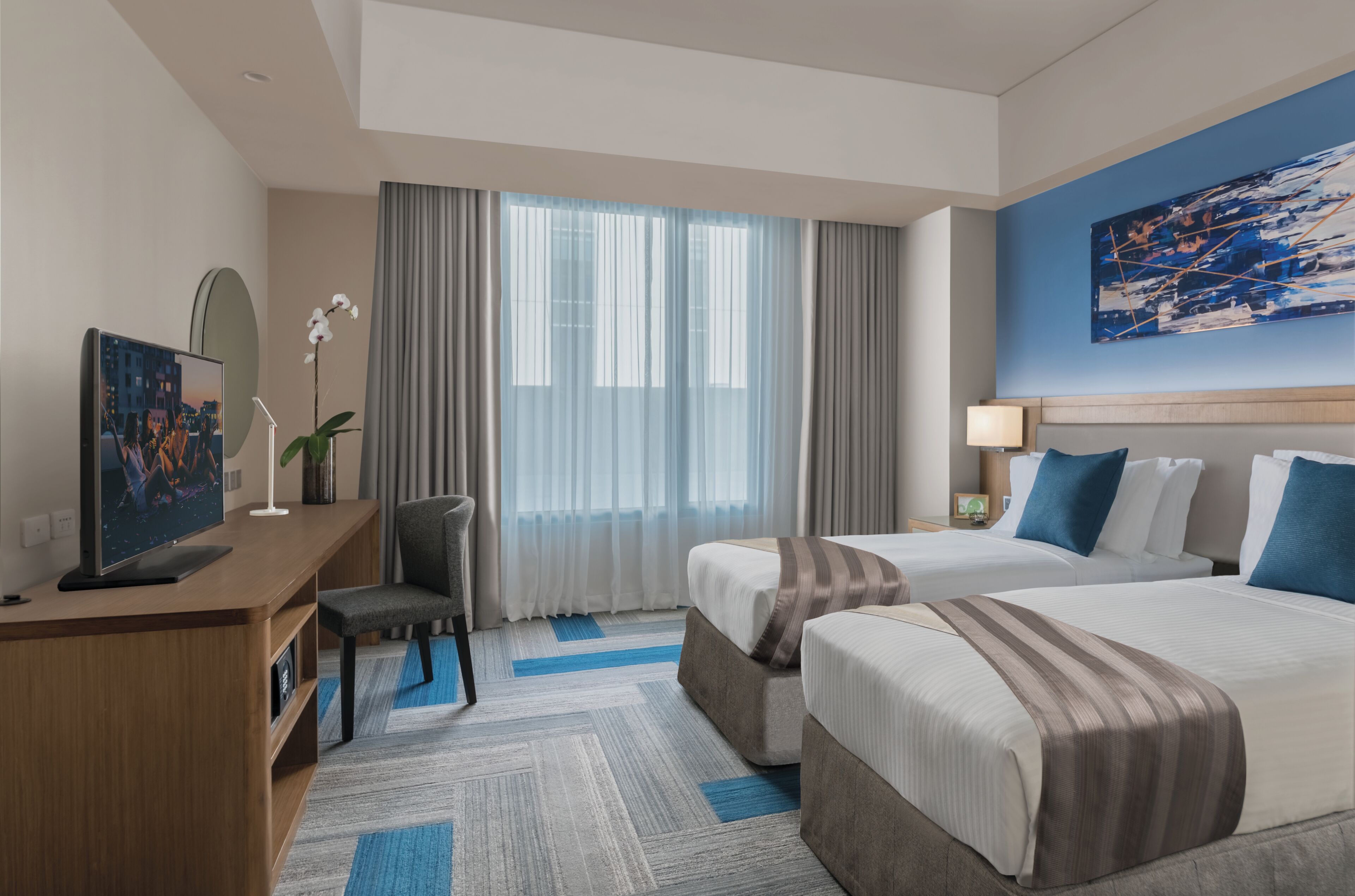 A hotel room with two beds, a desk with a TV, a chair, a lamp, and a painting on the wall.