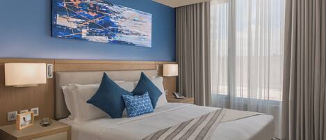 A hotel room with a large bed, two bedside tables, a nightstand, a painting on the wall, and a window with curtains.