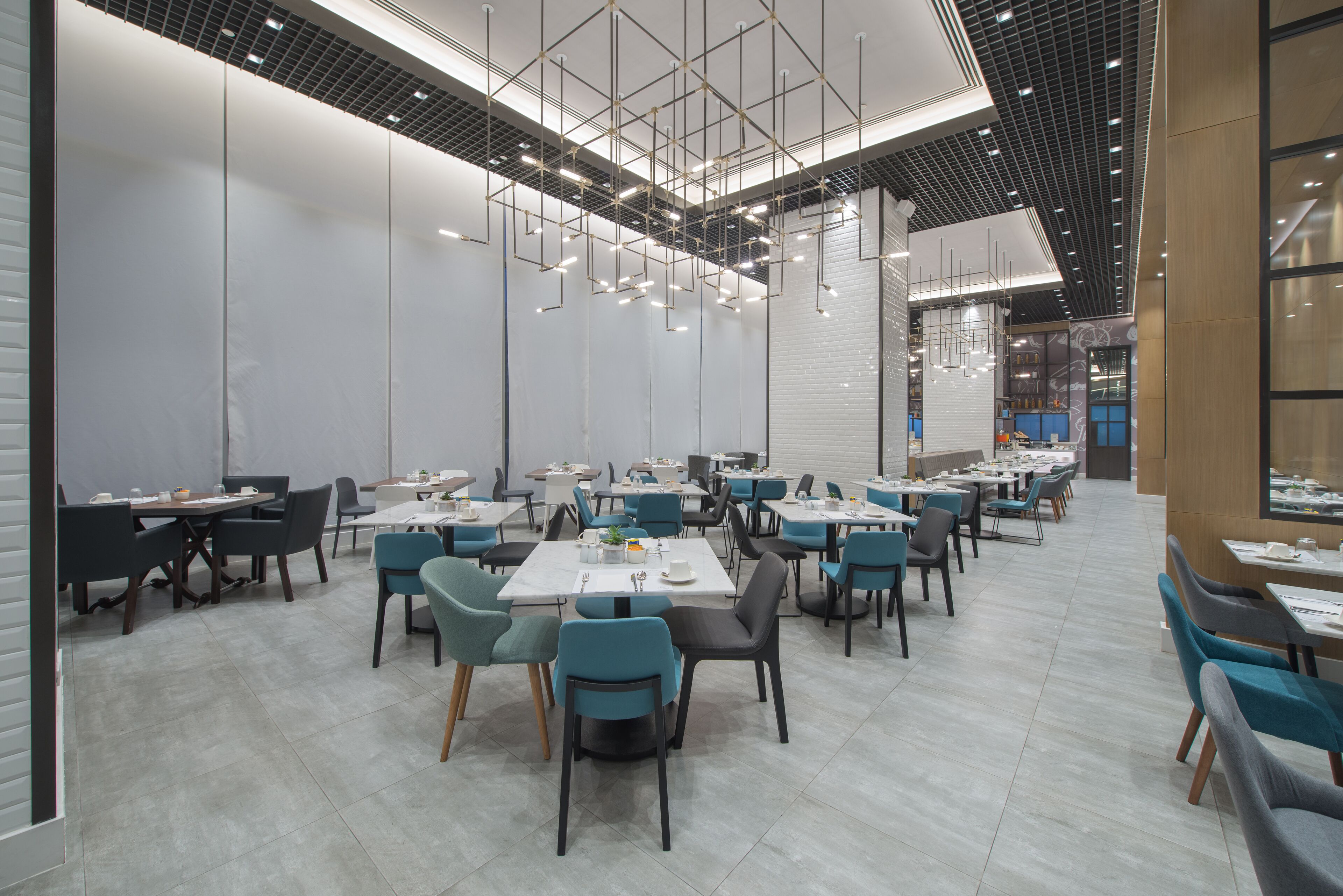 A modern dining area with multiple tables and chairs, a high ceiling with hanging lights, and a brick wall in the background.