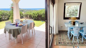 A dining area with a table set for two, a view of the sea, and a window with a view of greenery. - Dream holidays in Sardinia! (Chia)