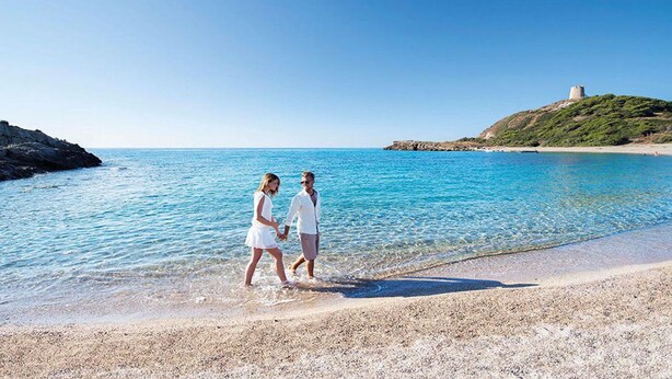 A couple walking on a beach with clear blue water and a castle on a hill in the background. - Dream holidays in Sardinia! (Chia)