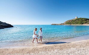 Ett par promenerar längs en strand med kristallklart blått vatten, och i bakgrunden syns ett slott på en kulle.