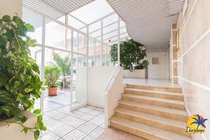 A spacious indoor area with a staircase leading to an upper level, surrounded by large windows and potted plants.
