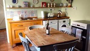 Una cocina con una mesa de madera, sillas, una lámpara colgante, una lavadora, una estufa y diversos utensilios de cocina y adornos en las paredes.
