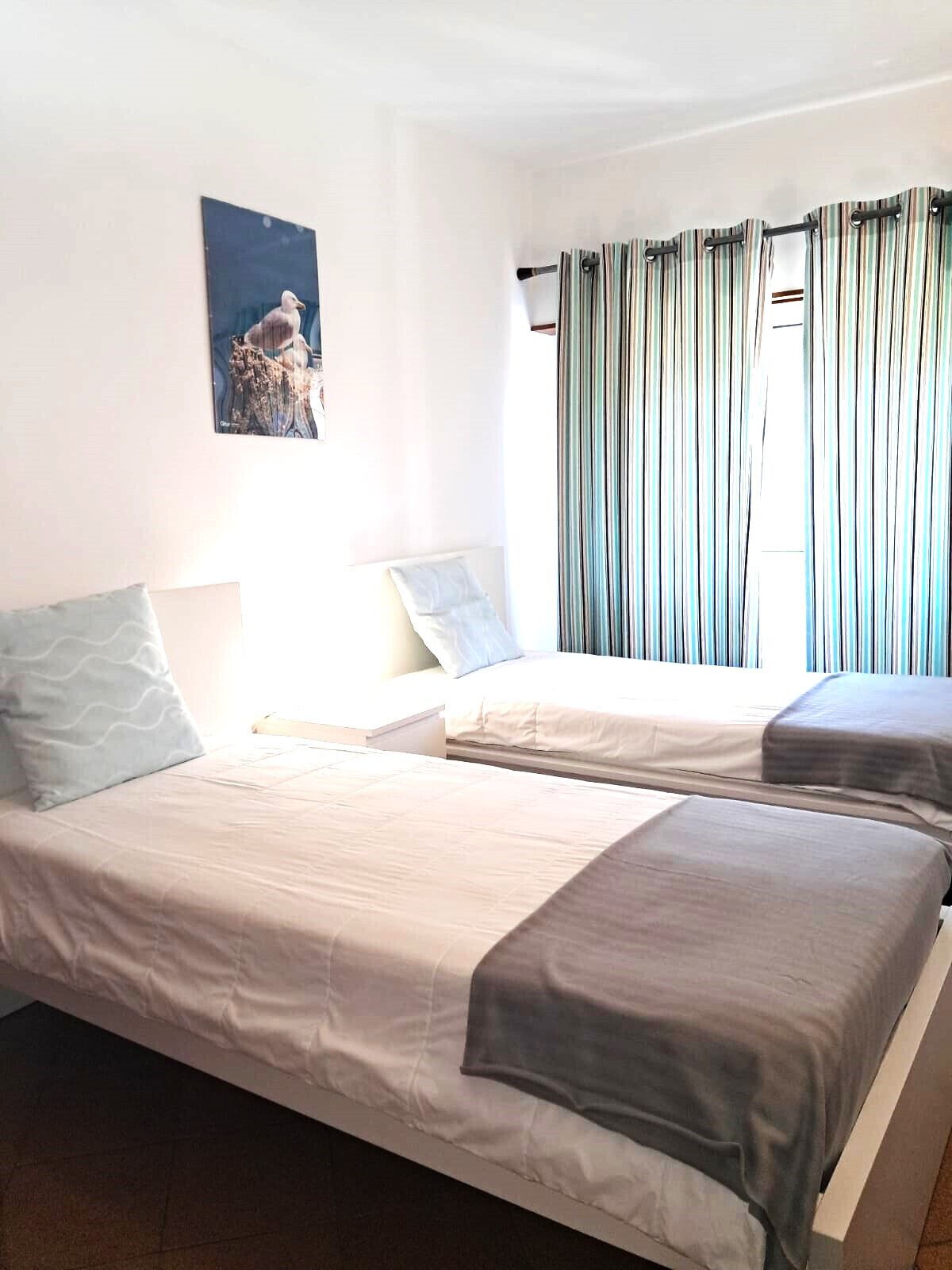 A room with two beds, a window with curtains, a painting on the wall, and a bedside table with a lamp.