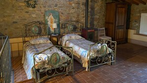 A rustic bedroom with two metal-framed beds, a stone wall, a wooden door, and a small table with a lamp.