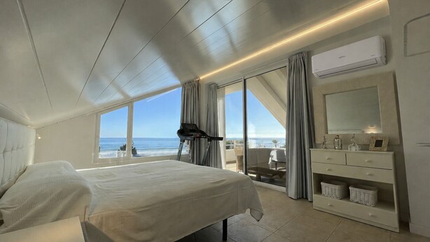 A bedroom with a bed, a treadmill, a large window with a view of the ocean, and a vanity with a mirror and drawers.
