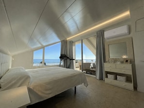 Une chambre à coucher comprenant un lit, un tapis de course, une grande fenêtre donnant sur l’océan, ainsi qu’une coiffeuse avec un miroir et des tiroirs.