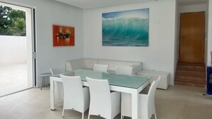 A dining area with a glass-top table, white wicker chairs, a white sofa, and a large wave painting on the wall.