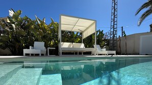 A clear swimming pool with tiled edges, surrounded by white lounge chairs and a covered seating area.