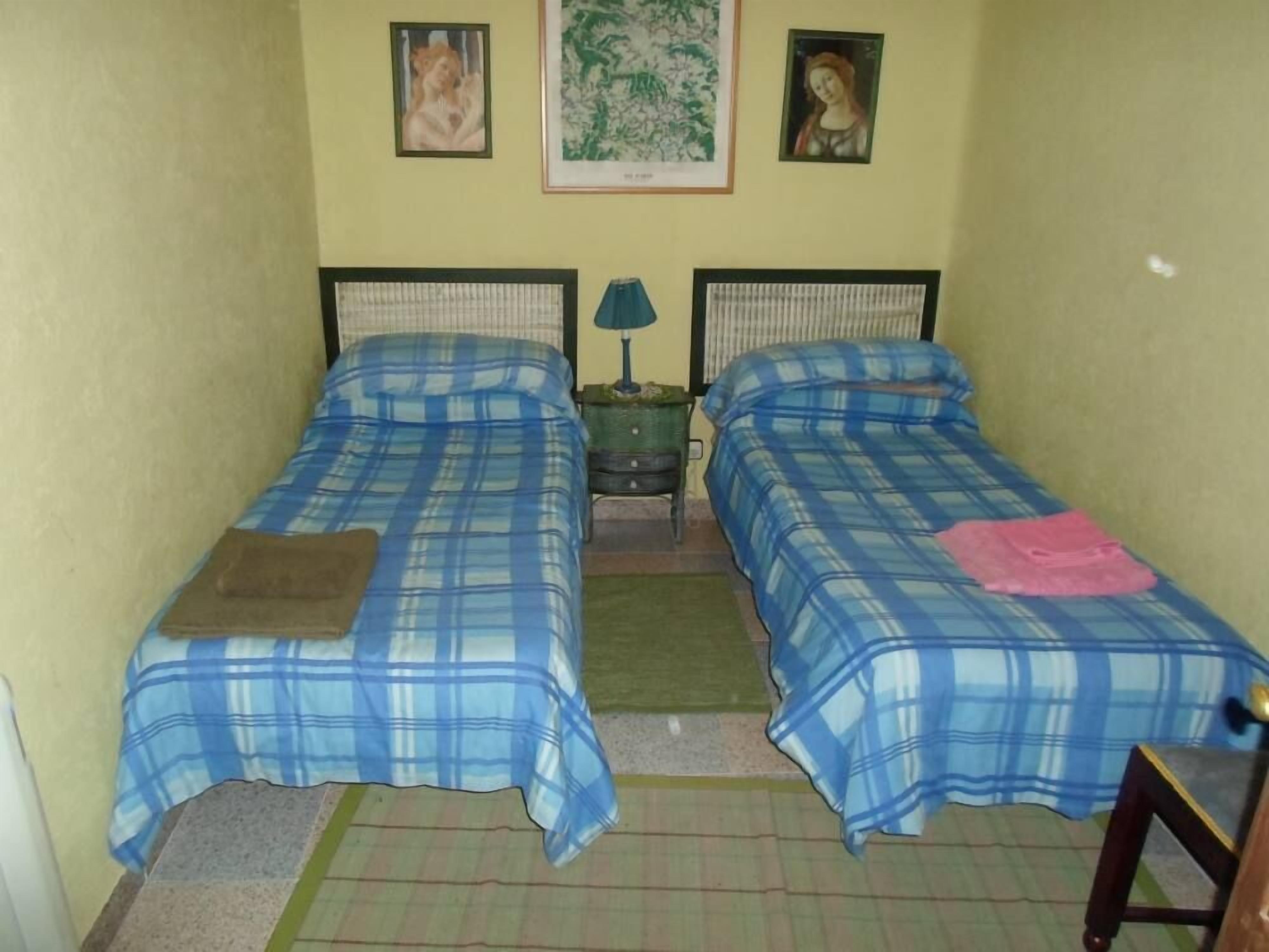 Una habitación pequeña con dos camas, cada una con ropa de cama a cuadros azules y blancos. Hay dos cuadros enmarcados en la pared, una mesita con una lámpara y un tapete verde en el suelo.