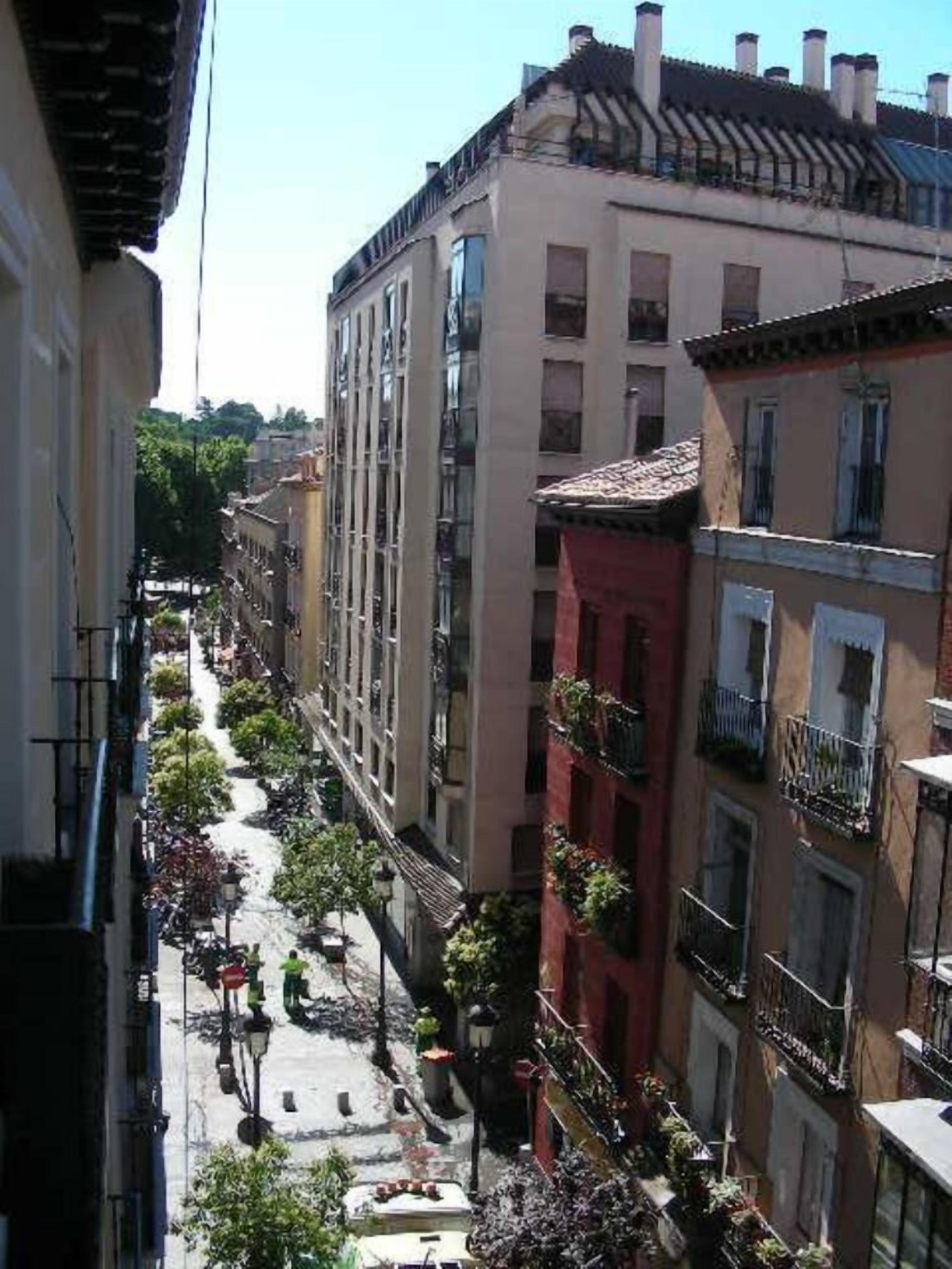 A narrow street lined with multi-story buildings, featuring balconies with plants and a central pedestrian area with street lamps.