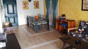 Ein Wohnzimmer mit Essbereich, einer Couch, einem Tisch und einem Stuhl. Eine Tür, ein Fenster mit Vorhängen und eine Wand mit Kunstwerken sind zu sehen.