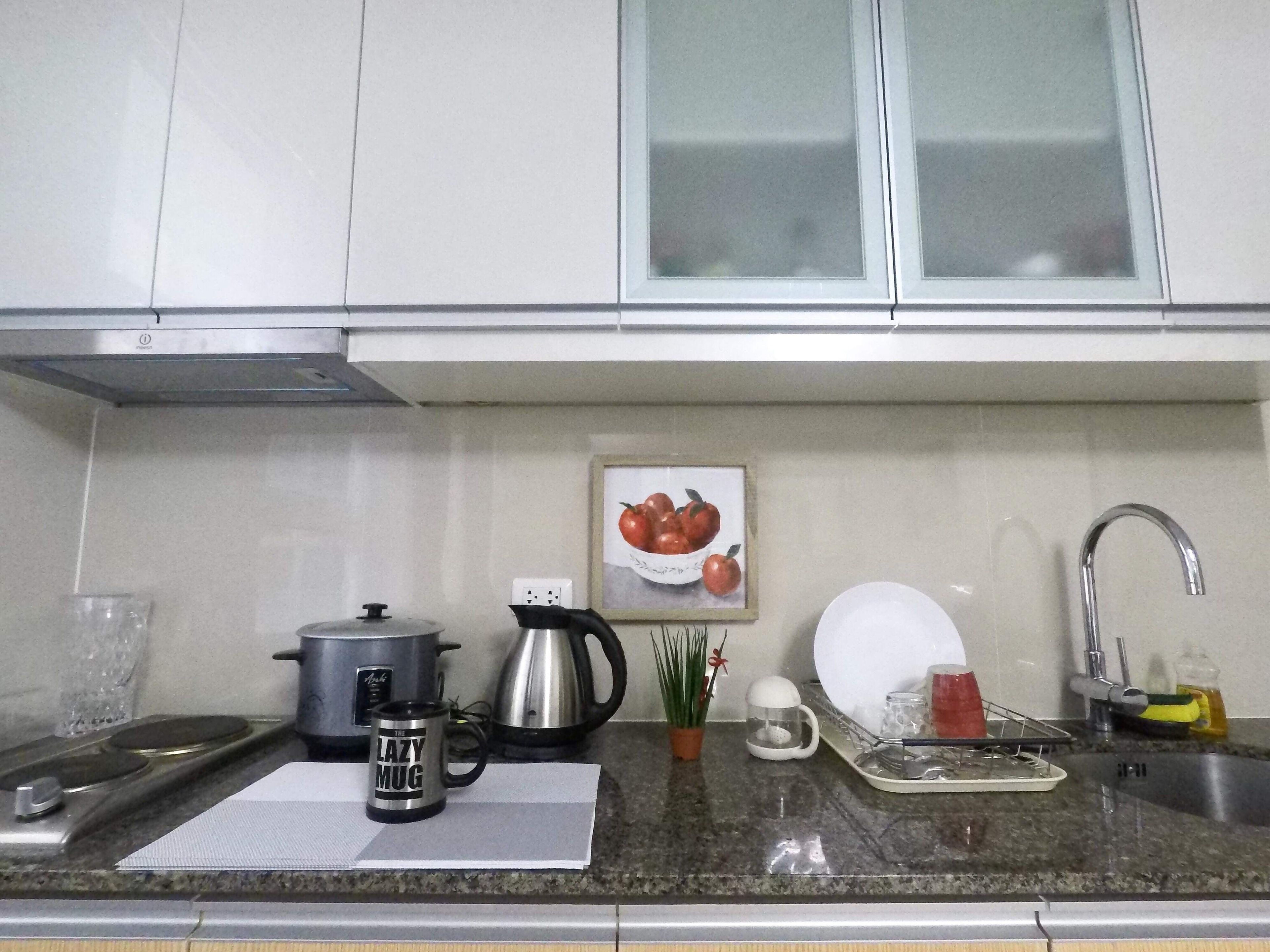 A kitchen with white cabinets, a stainless steel kettle, a coffee maker, a potted plant, and a framed picture of apples.