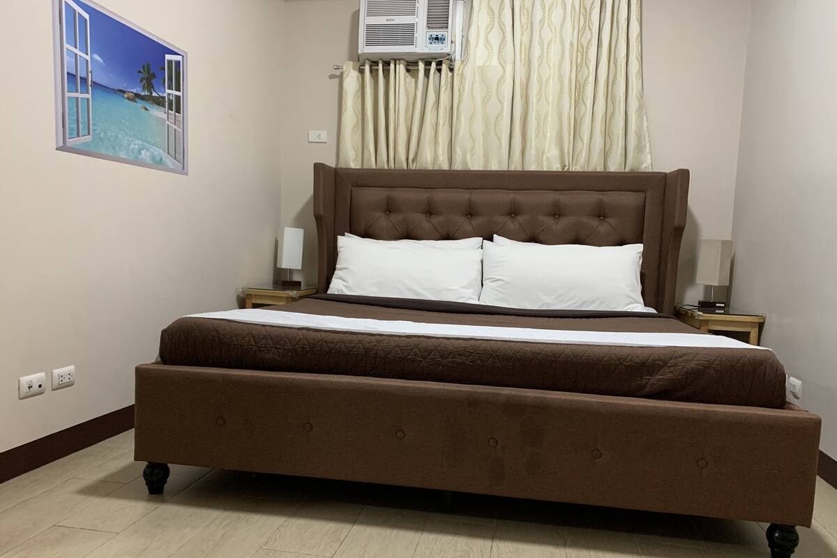 A large bed with a brown headboard, two white pillows, and a brown bedspread. There is a nightstand with a lamp and a wall-mounted air conditioner.