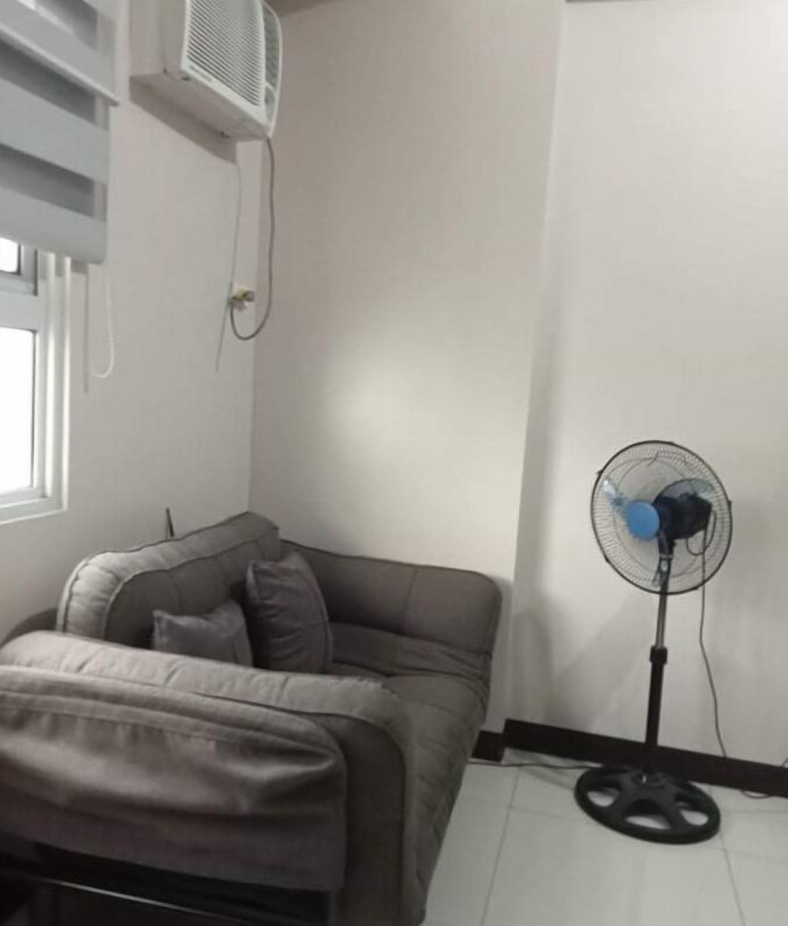 A room with a grey sofa, a standing fan, and an air conditioning unit on the wall.