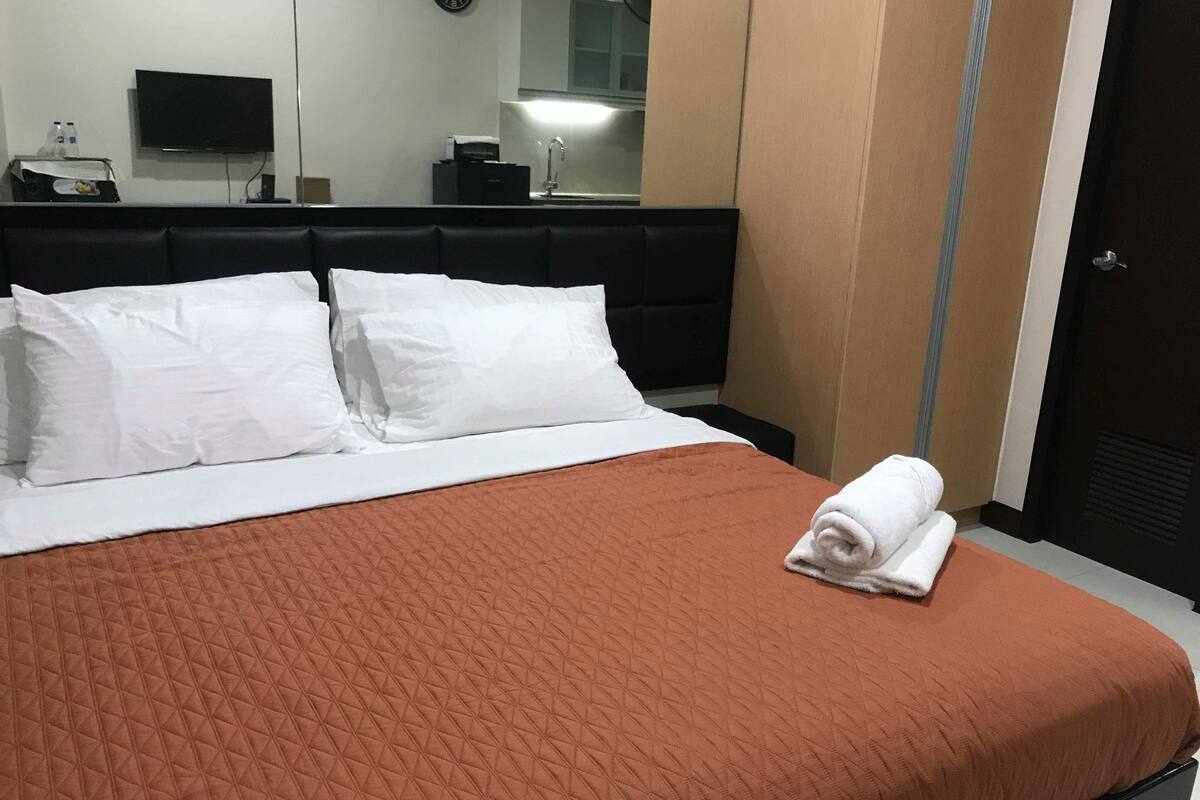 A hotel room with a bed, white pillows, a brown quilted bedspread, a folded towel, a flat-screen TV, a clock, and a mirror.