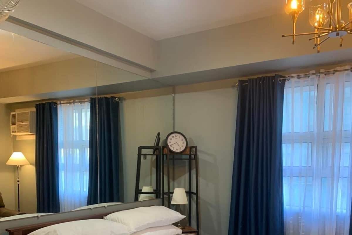 A neatly made bed with white and blue bedding, a bedside lamp, a clock on the wall, and a ceiling fan.