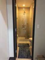 Corredor estreito que dá para um espaço com chuveiro, porta de vidro, sanita e um tapete pequeno no chão.