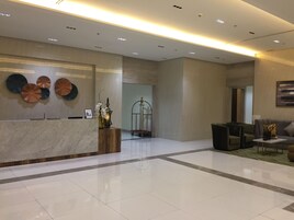 Um lobby moderno com uma receção de mármore, uma parede decorativa com placas de metal circulares, um portão em estrutura de metal e uma área de estar com sofás e poltronas.