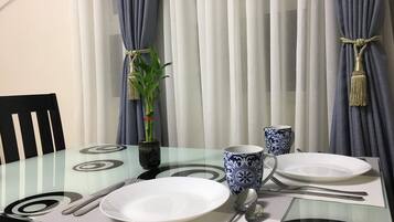 Una mesa de comedor dispuesta con platos blancos, cubiertos y una taza con diseño azul y blanco. Hay una planta en maceta sobre la mesa, y la habitación tiene cortinas con flecos.