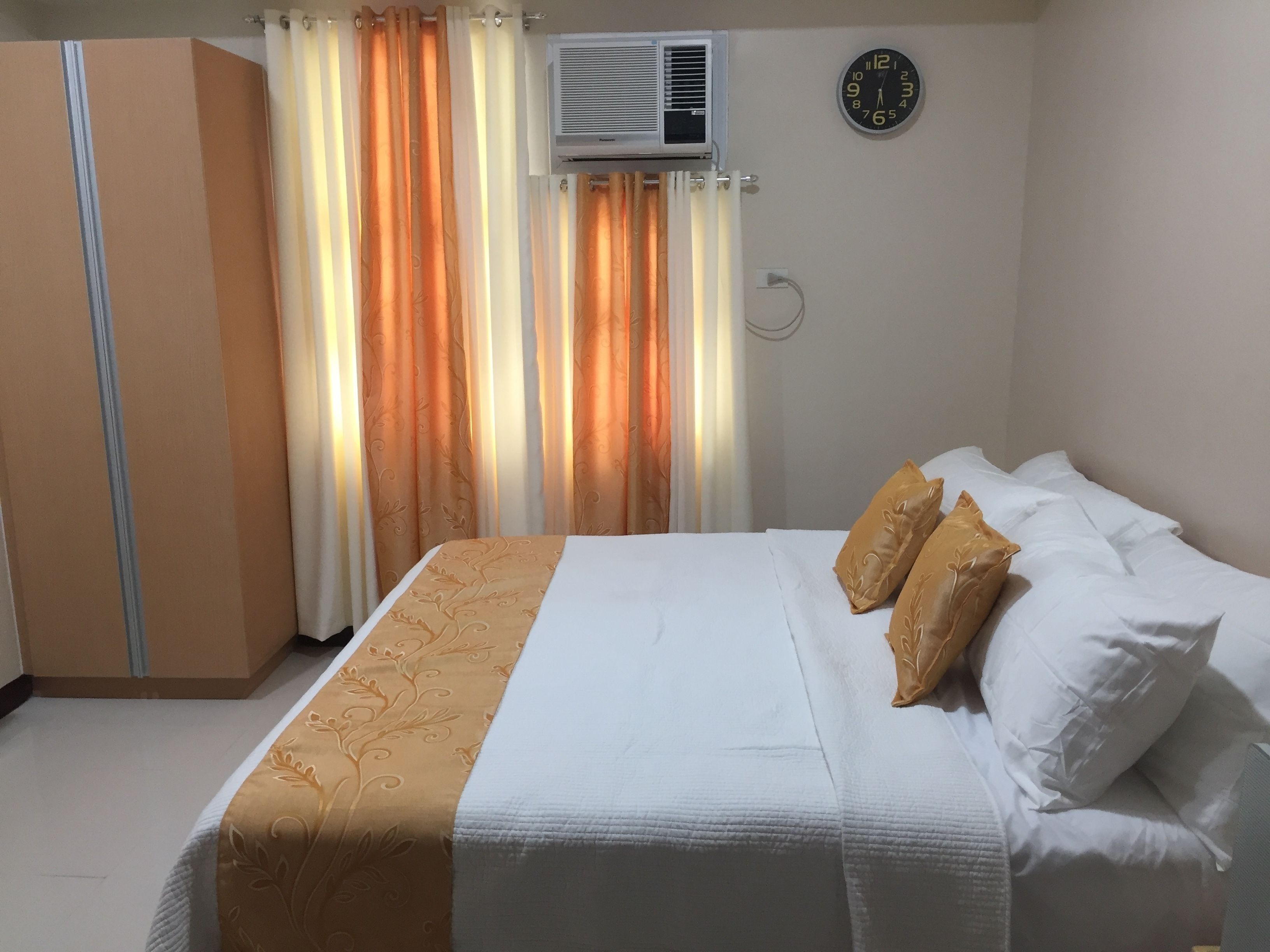 A bedroom with a bed, white pillows, a beige and white bedspread, an air conditioning unit, a clock, and curtains.