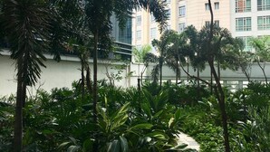 Un patio con abundante vegetación, un camino y edificios modernos con una torre del reloj al fondo.