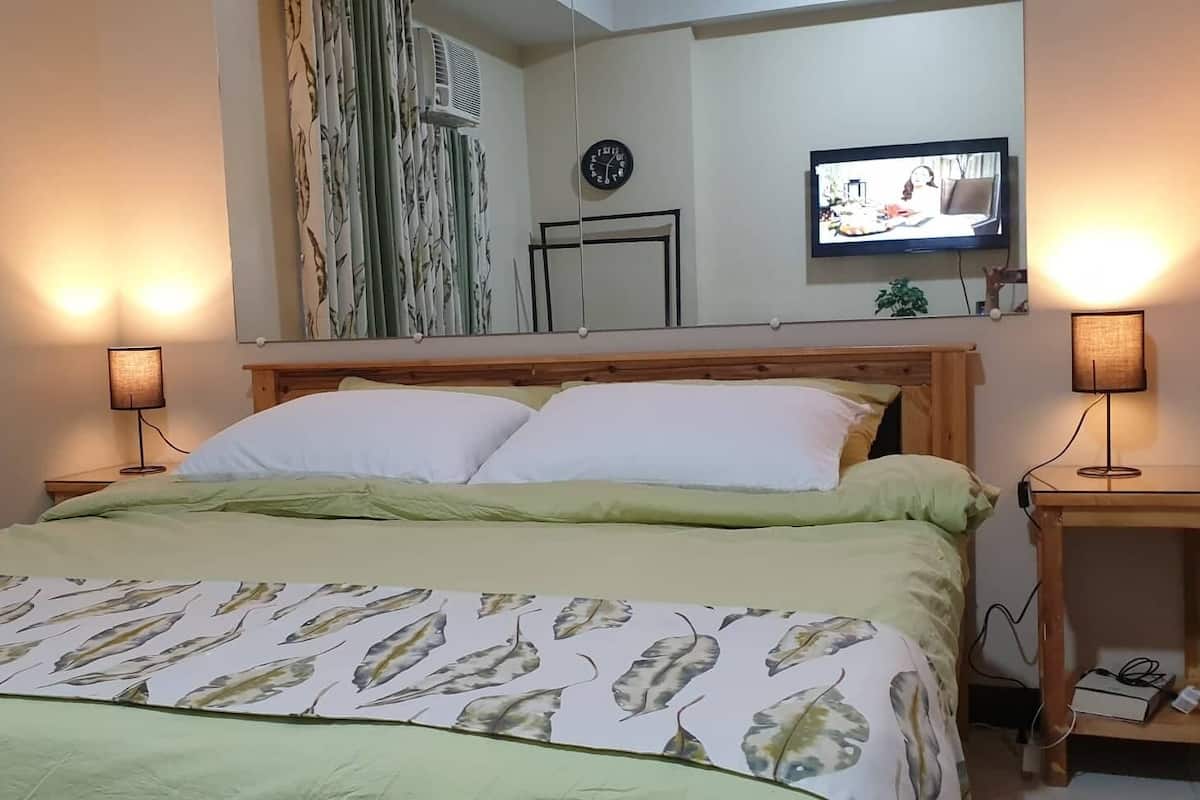 A neatly made bed with a green and white leaf-patterned bedspread, two bedside lamps, a clock, and a television on the wall.