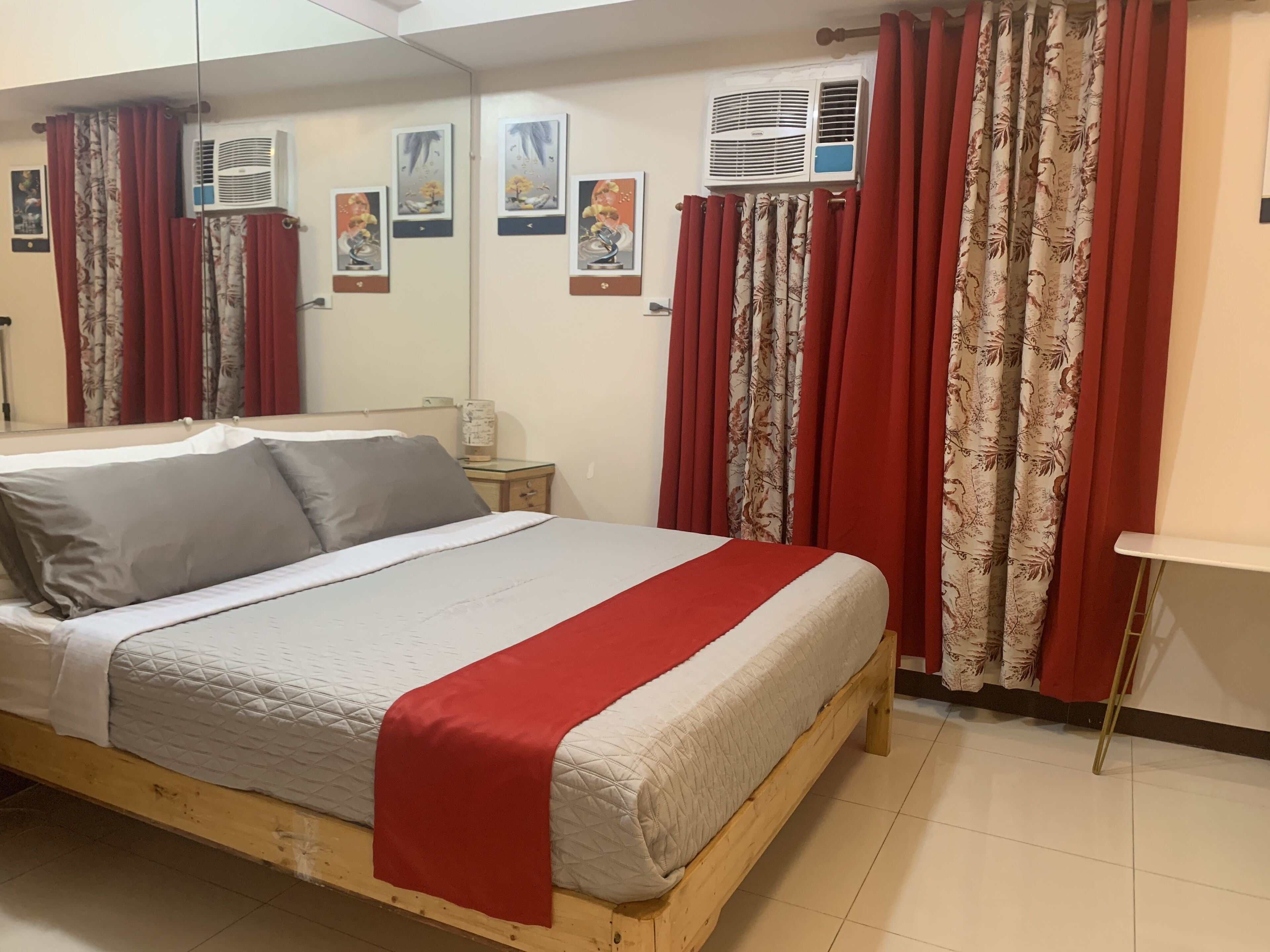 Un dormitorio con una cama de madera, cortinas rojas con estampado floral, unidades de aire acondicionado y cuadros en la pared.