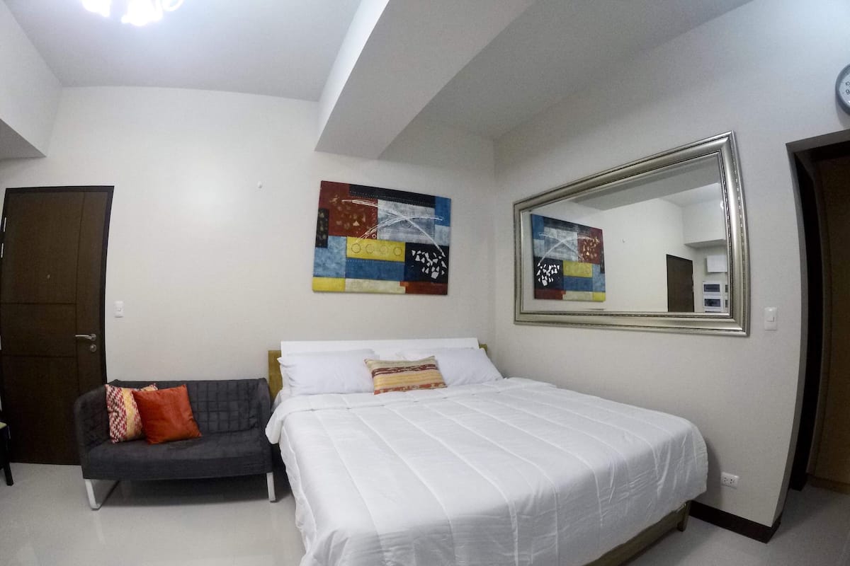 A bedroom with a bed, a sofa, a colorful abstract painting, and a mirror.