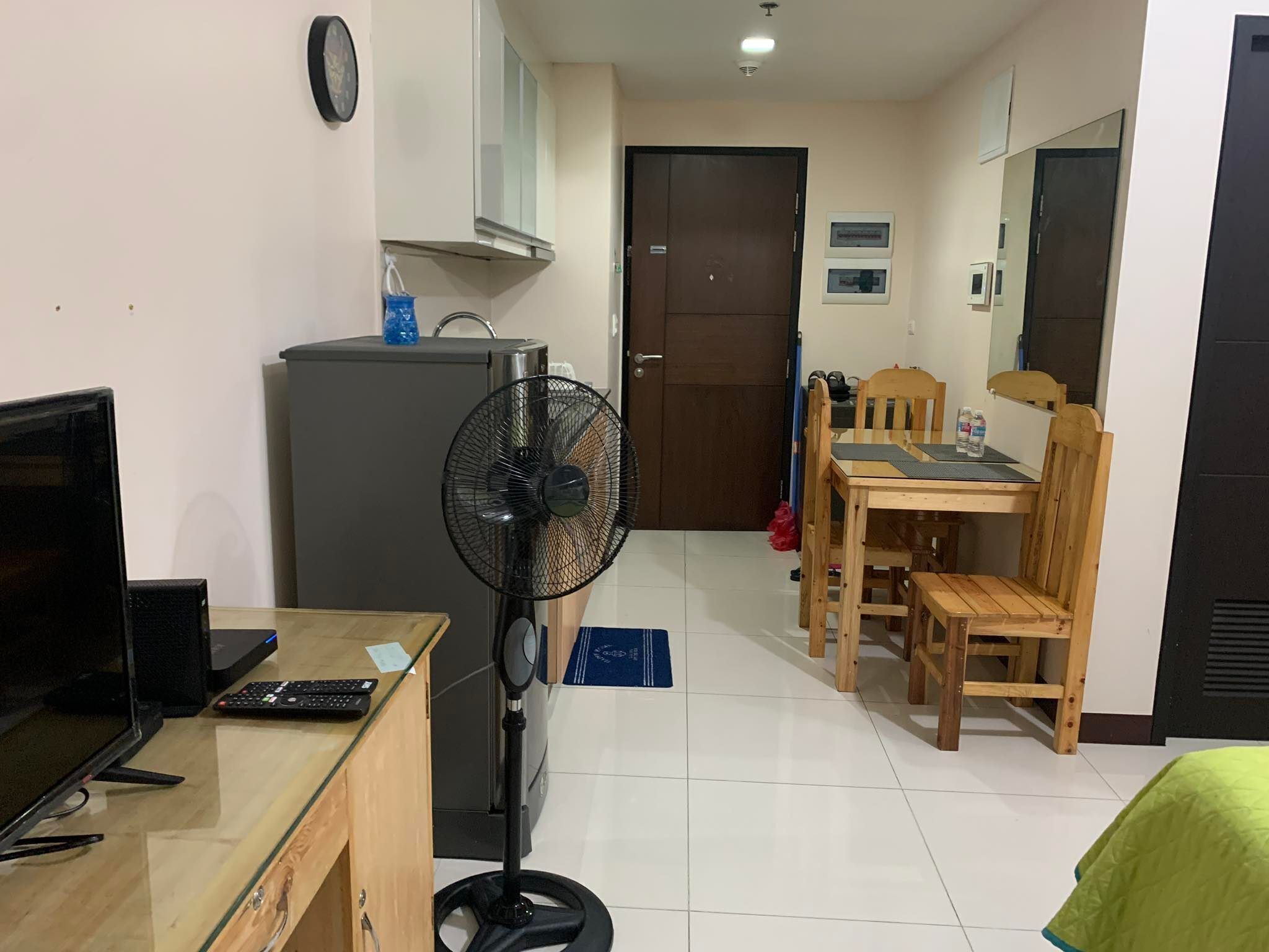 Une pièce de taille modeste comprenant un bureau, une chaise, une table à manger avec des chaises, un réfrigérateur, un ventilateur et un lit recouvert d’une housse verte.