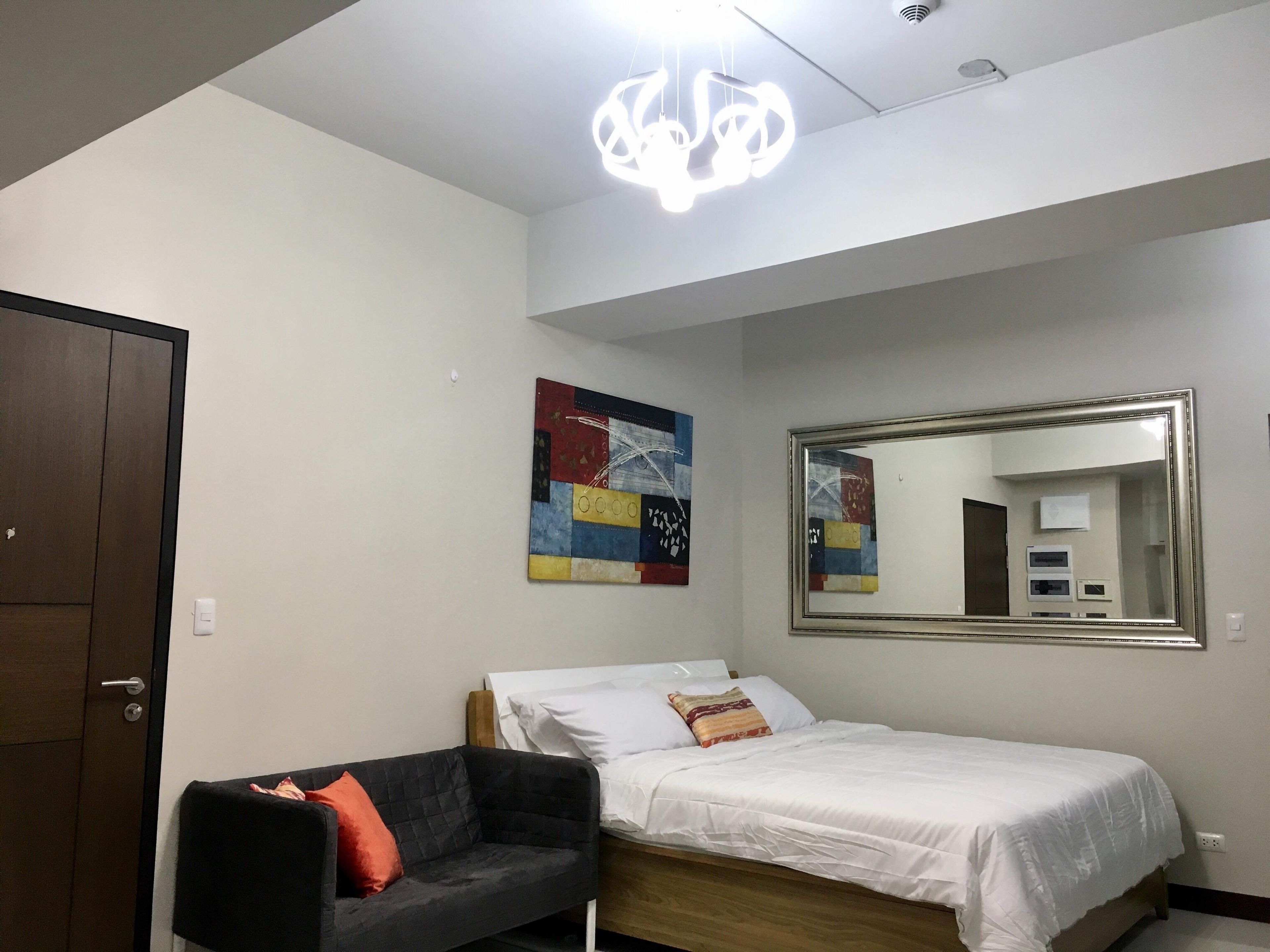 A bedroom with a bed, a black couch, a colorful abstract painting, a large mirror, and a ceiling light fixture.