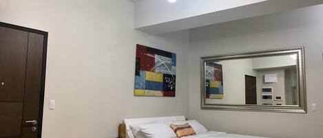 A bedroom with a bed, a black couch, a colorful abstract painting, a large mirror, and a ceiling light fixture.