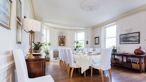 Uma sala de jantar com uma mesa comprida posta para refeição, rodeada por cadeiras brancas, piso de madeira e diversos objetos decorativos.