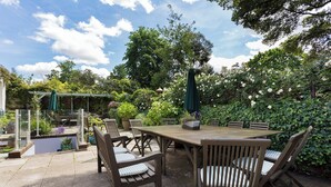 Um pátio externo com mesa e cadeiras de madeira, rodeado por vegetação exuberante e plantas floridas.