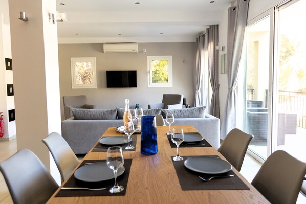 A modern dining area with a wooden table set for four, grey sofas, and a TV mounted on the wall.