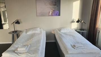 Quarto de hotel com duas camas de solteiro, cada uma com roupa de cama e almofadas brancas, uma mesa redonda com um abajur e um quadro na parede.