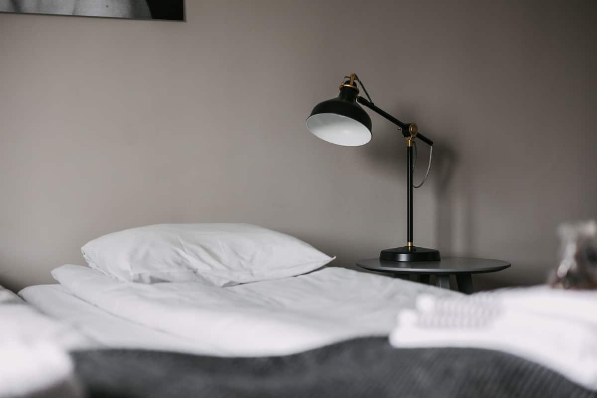 A neatly made bed with white sheets and pillows, a round black side table with a black desk lamp, and a plain wall in the background.