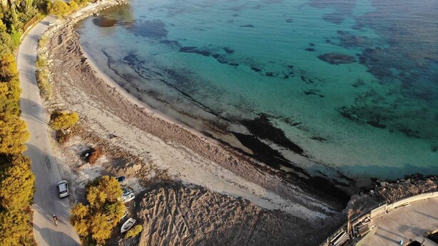 Eine Luftaufnahme einer Küstenregion mit einer sich an der Uferlinie schlängelnden Straße, einem Strand und einer kleinen Siedlung mit Gebäuden und Bäumen.