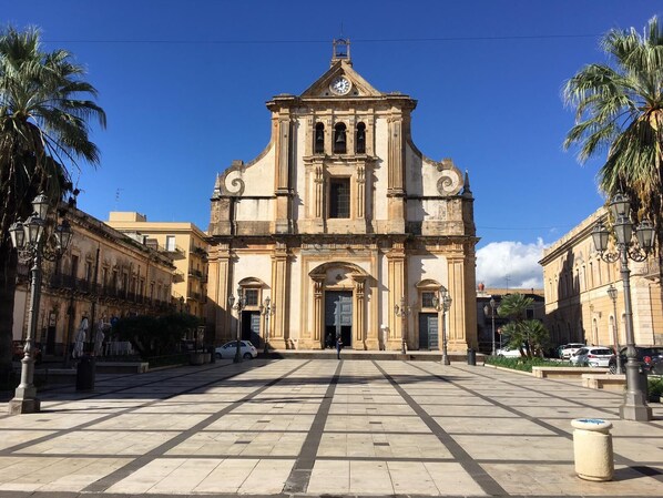 A historic building with a central entrance, flanked by palm trees and a paved square.