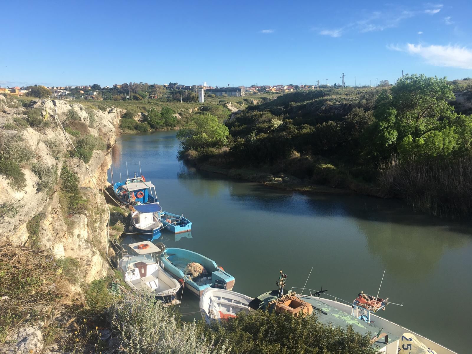 Une rivière bordée de bateaux amarrés, entourée de verdure et avec une ville visible au loin.